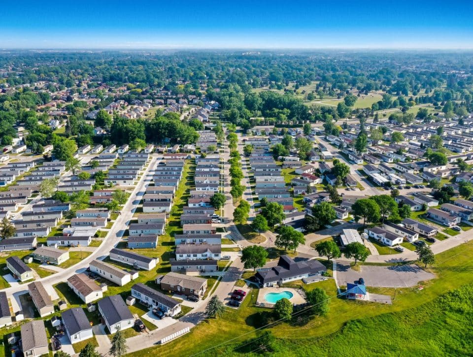 Aerial view of Rivers Edge, a suburban neighborhood with rows of houses, green lawns, trees, and a community swimming pool, stretching towards a distant horizon under a clear blue sky.