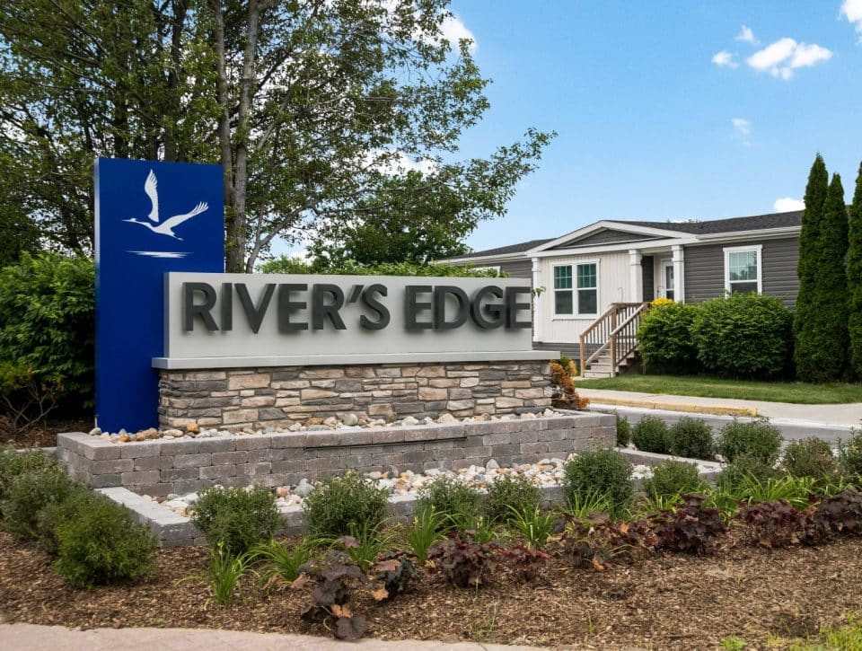 A stone and concrete sign reading Rivers Edge stands in landscaped greenery and small shrubs, with a house and tall trees in the background under a blue sky with clouds.