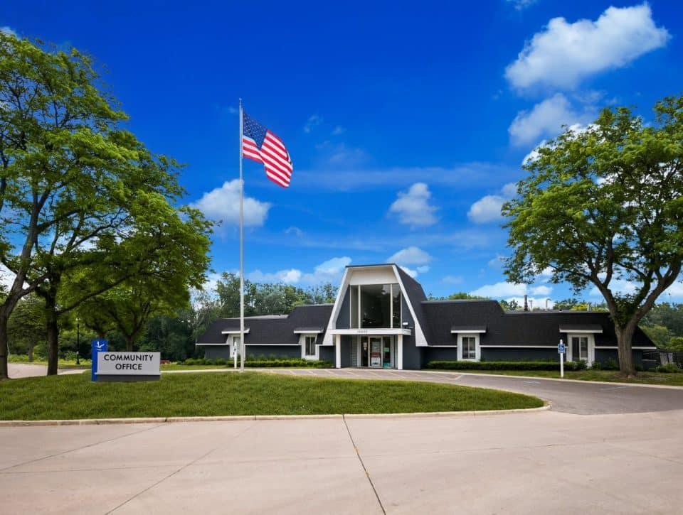 A modern community office building with large windows stands at Rivers Edge, surrounded by trees and grass under a bright blue sky. An American flag waves near the entrance of this all age MH community with great amenities.