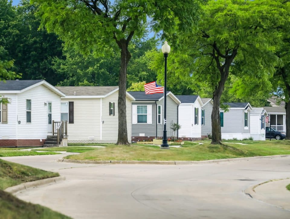 A row of affordable homes lines a curved street with green lawns and leafy trees in Clinton Township. An American flag hangs on a lamppost, and several cars are parked beside the homes on a sunny day.