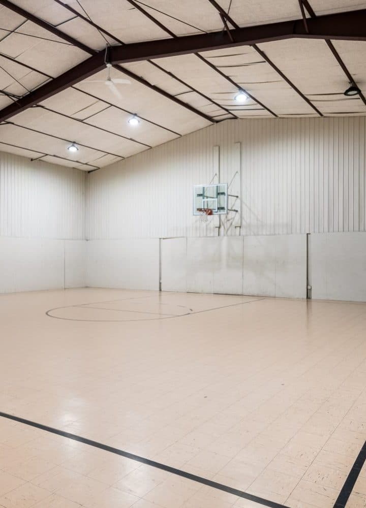 An empty indoor basketball court at Rivers Edge features a wooden floor, white walls, a basketball hoop, and overhead lights. The high-ceilinged space has exposed beams and a closed door at the back.