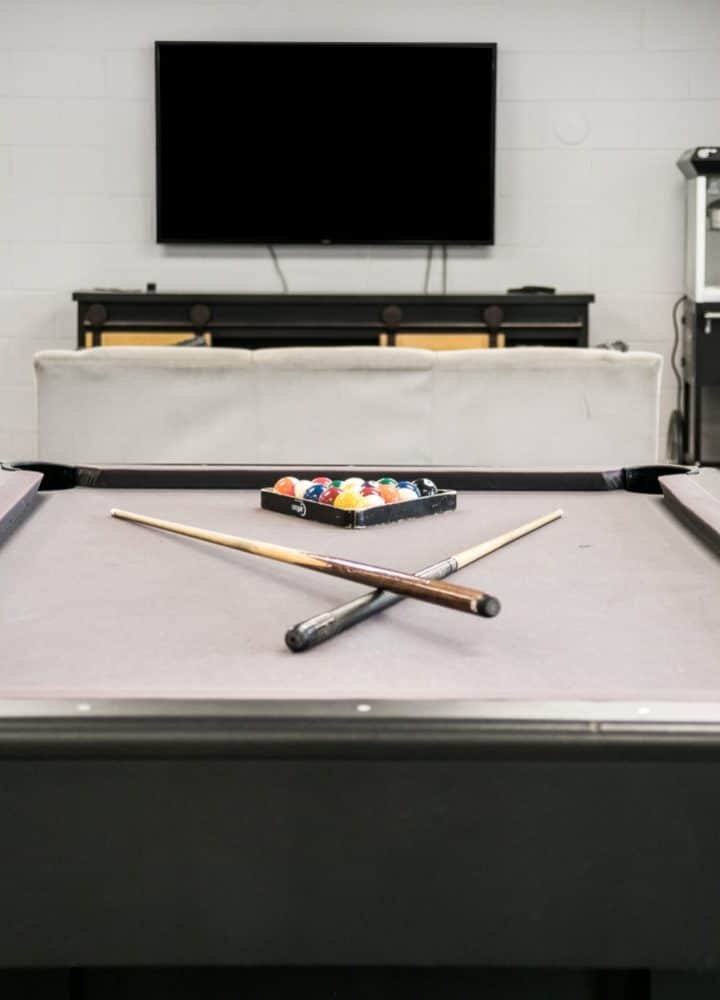 A modern game room at Rivers Edge features a pool table with two cues crossed and racked balls, a sofa, two upholstered chairs, a TV mounted on the wall, and a popcorn machine in the background.