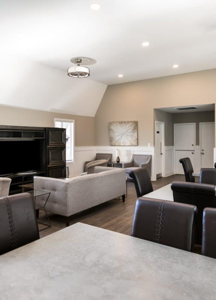A modern, well-lit lounge at Rivers Edge features a TV, gray couches, dark wood furniture, and multiple tables with black chairs. Light beige and white walls are adorned with framed artwork and contemporary lighting.