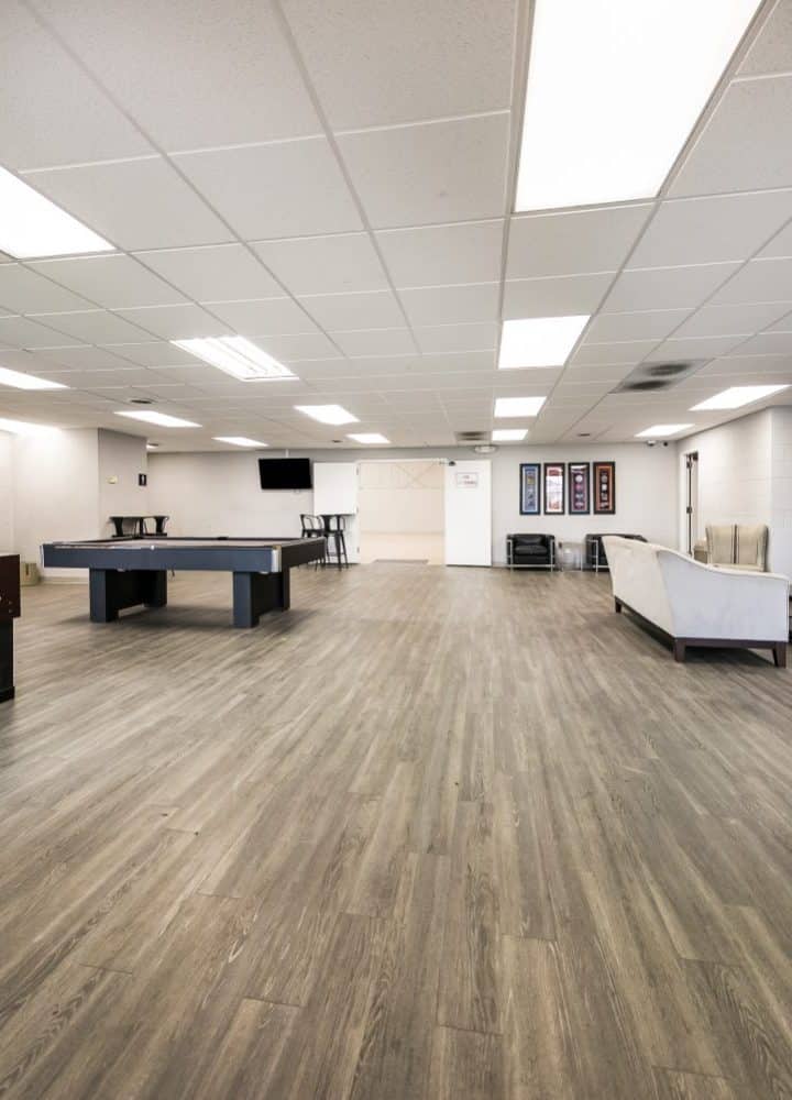 Spacious recreation room at Rivers Edge with foosball and pool tables, modern sofas, chairs, flat-screen TVs on the walls, and wood-style flooring under bright ceiling lights.