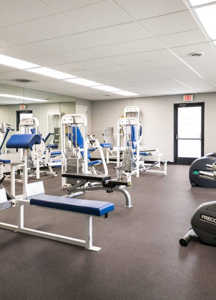 A bright gym room at Rivers Edge features various exercise equipment, including weight machines, benches, stationary bikes, and elliptical trainers, all neatly arranged on a dark floor under fluorescent lighting.