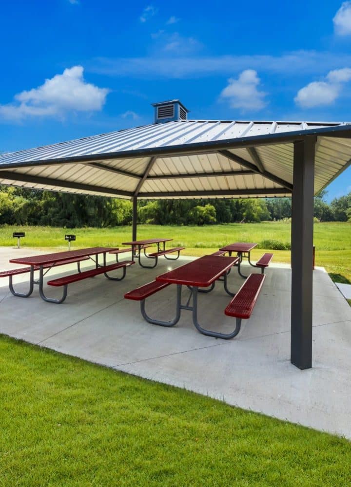 A park pavilion with a metal roof shelters several red picnic tables on a concrete pad, surrounded by green grass, trees, and a trash can—one of the great amenities offered in this all age MH community under a sunny blue sky with scattered clouds.