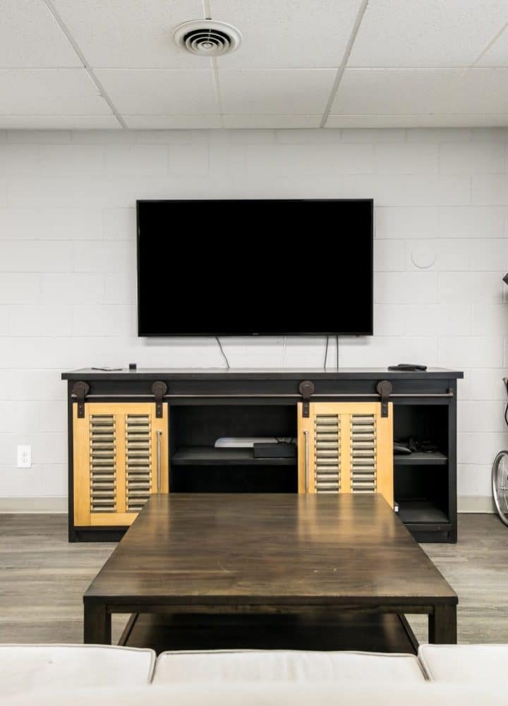 A modern lounge at Rivers Edge features a wall-mounted TV, dark wooden console, popcorn machine on a cart, wooden coffee table, and light-colored sofas arranged around it. White walls and a wood floor complete the inviting atmosphere.