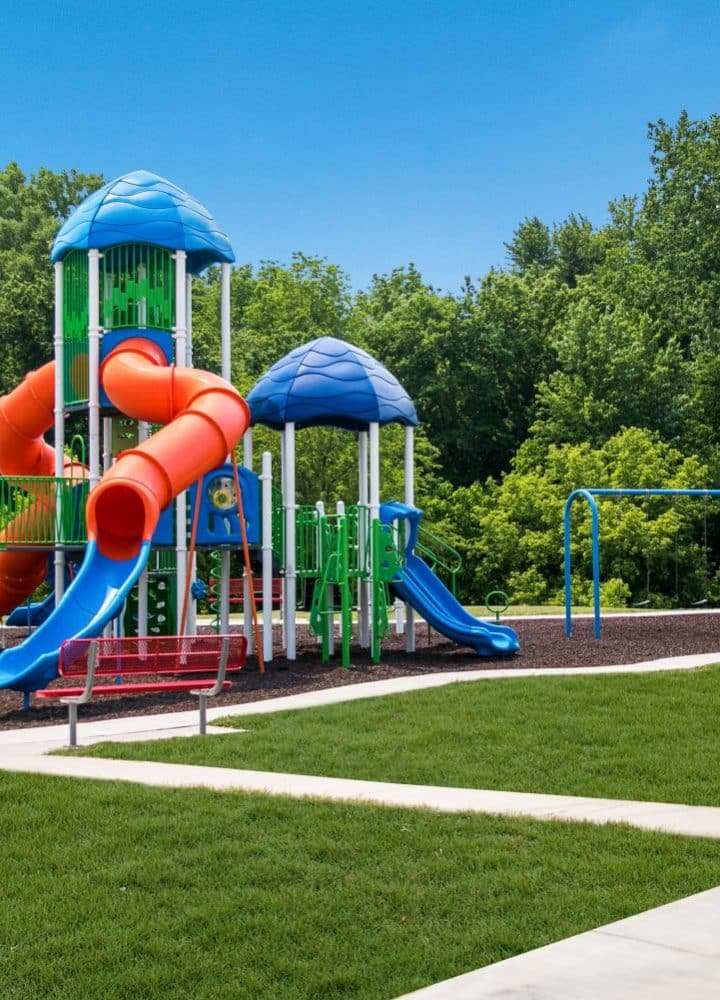 A colorful playground at Rivers Edge in Clinton Township features blue and orange slides, green climbing structures, and blue swings on mulch, all surrounded by grass and trees under a clear sky in this all age MH community.
