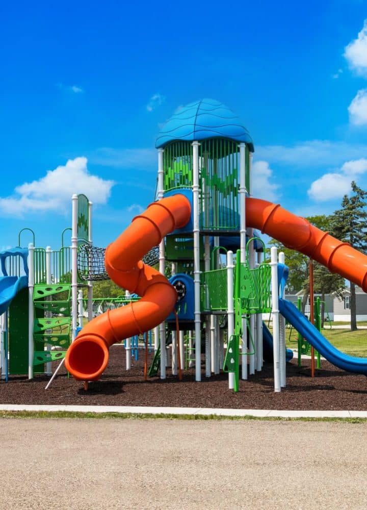A colorful outdoor playground at Rivers Edge features tall climbing structures, bright orange tube slides, green and blue platforms, and surrounding benches, all set on a grassy area under a blue sky with scattered clouds.