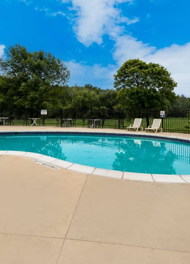 Outdoor swimming pool at Rivers Edge, surrounded by lounge chairs and tables, enclosed by a black metal fence, with green trees and a bright blue sky with clouds in the background.