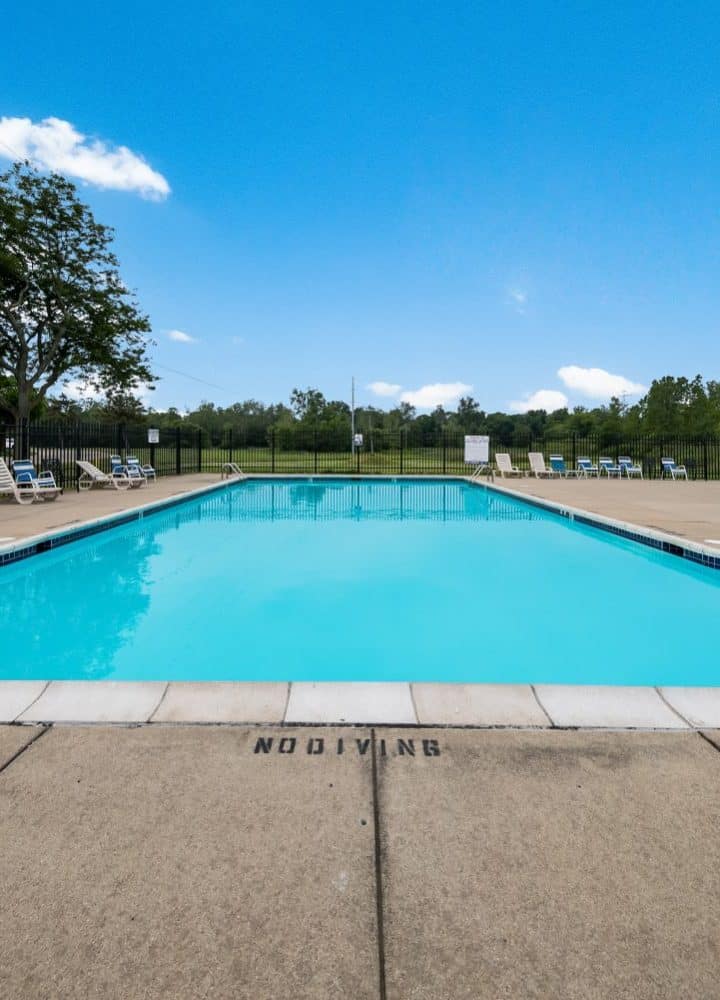 An outdoor swimming pool with clear blue water sits at Rivers Edge, surrounded by lounge chairs and trees under a bright blue sky. The pool deck is empty, and a No Diving warning is visible on the edge.