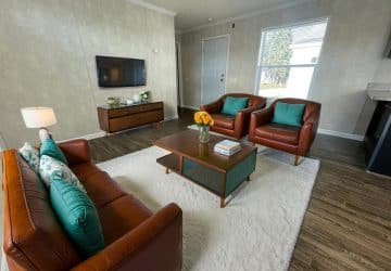 A modern living room with brown leather sofa and chairs, teal accent pillows, a wooden coffee table, TV on the wall, and a white rug on wooden floors. A window lets in natural light near a white front door.