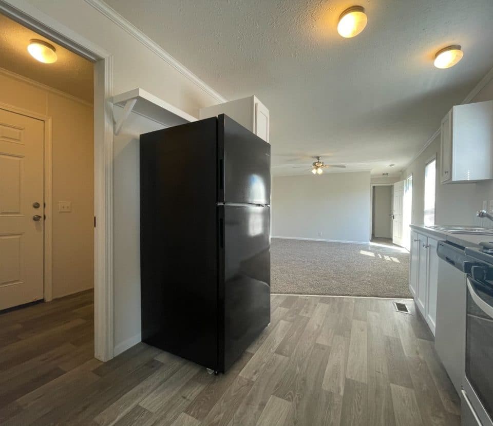 A small, modern kitchen with light wood flooring, a black refrigerator, white cabinets, and a stove. The kitchen opens into a carpeted living area with ceiling fan and several windows letting in natural light.