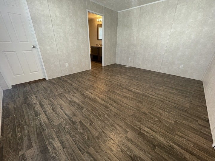 Empty room with light-colored textured walls, dark wood flooring, a white door on the left, and an open doorway leading to a bathroom with a lit vanity mirror in the background.
