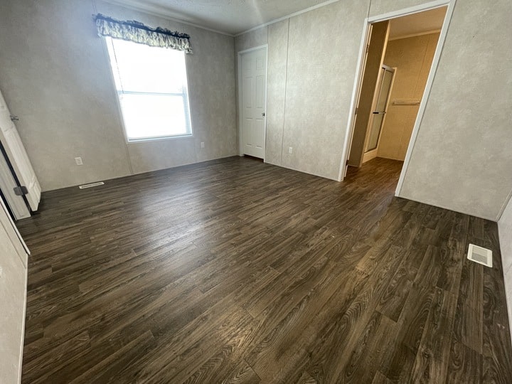 A spacious, empty room with wood-look flooring, light-colored walls, a large window with a floral valance, a closed white door, and an open doorway leading to a bathroom with a visible shower.