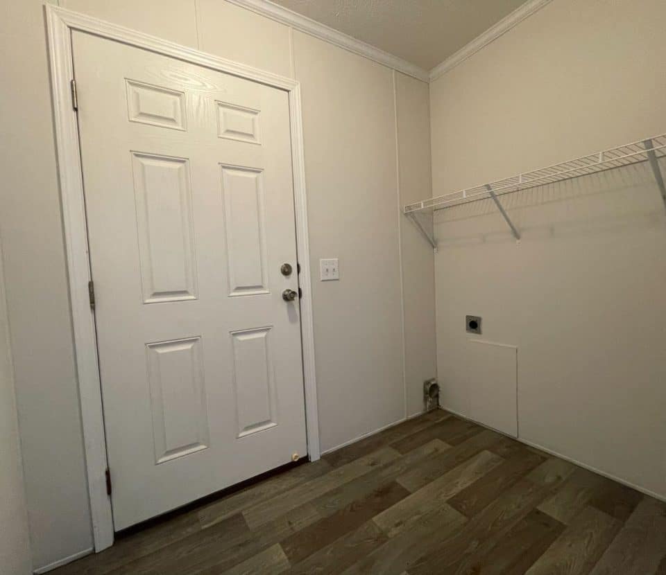 A small, empty laundry room with a white door, beige walls, wood-look flooring, a white wire shelf, a fire extinguisher, and wall hookups for a washer and dryer.