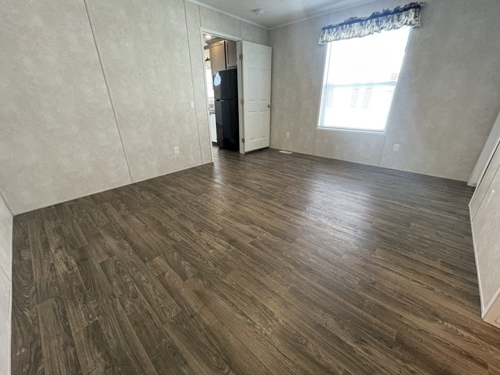 An empty room with wood-style flooring, light-colored walls, a large window with a curtain, and a white door leading to a kitchen area with visible appliances.