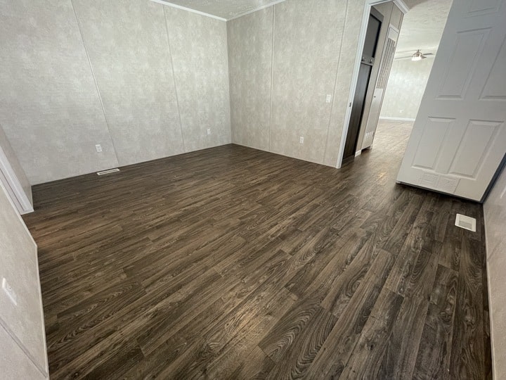 Empty room with dark wood flooring, light-colored textured walls, baseboards, white electrical outlets, and open doorways leading to adjacent rooms. Ceiling fan is visible in the background.