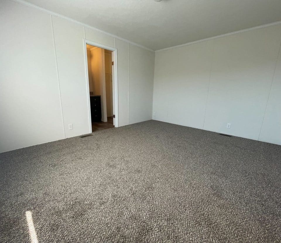Empty room with light beige walls, carpeted floor, an open doorway leading to another area, and sunlight casting a strip of light on the carpet near the bottom left corner.