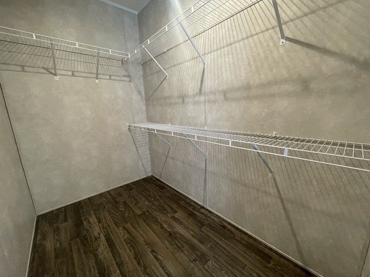 Empty walk-in closet with light beige walls, dark wood flooring, and white wire shelves mounted on two walls at different heights. The space is well-lit and uncluttered.
