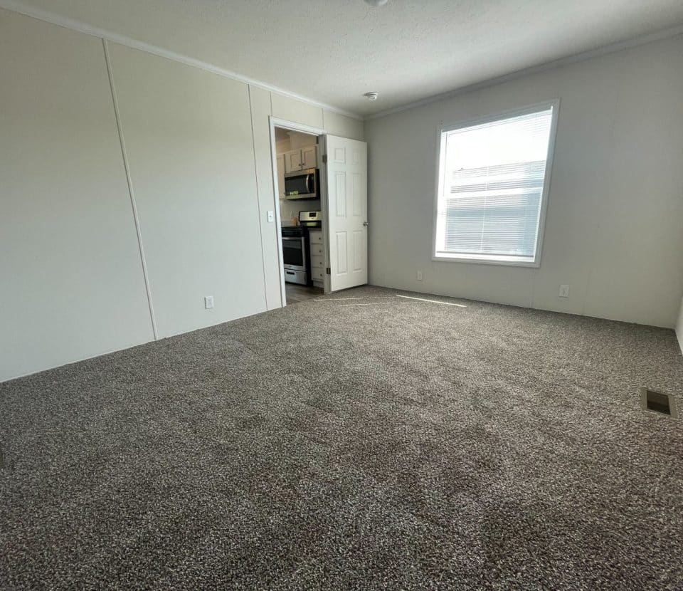 A spacious, empty room with light gray carpet, white walls, a large window letting in natural light, and an open doorway leading to a kitchen with white cabinets and appliances.