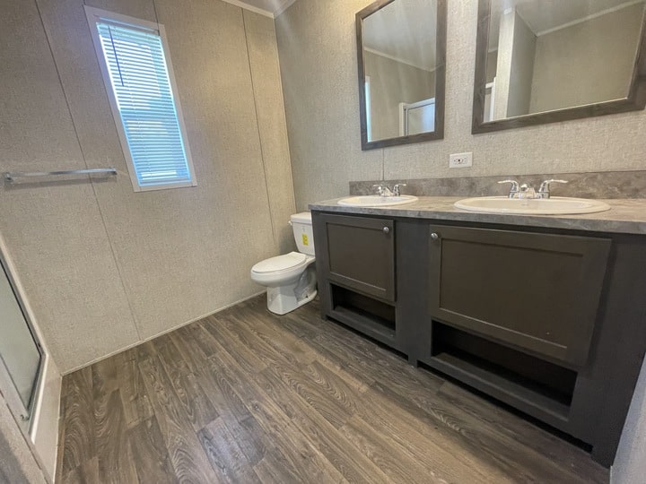 A modern bathroom with wood-patterned flooring, a toilet, a glass shower, a double-sink vanity with dark cabinets, two mirrors above the sinks, and a window with blinds on the left wall.