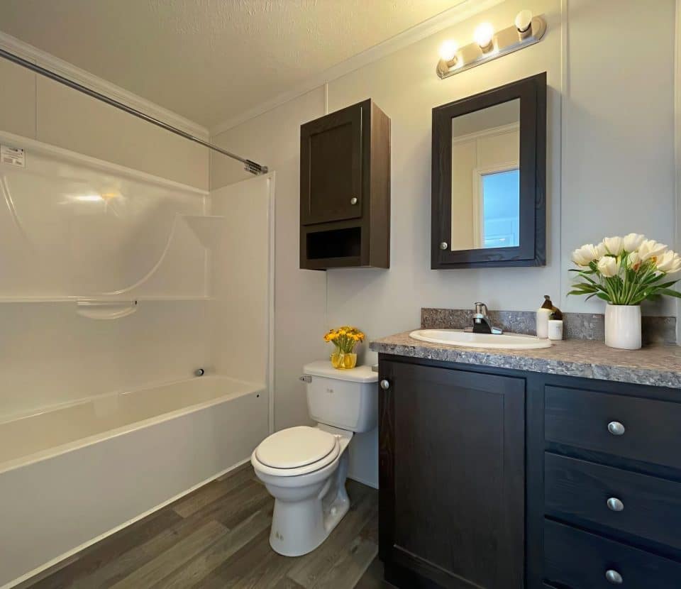 Modern bathroom with a bathtub-shower combo, toilet, and dark wood vanity. A mirror cabinet is above the sink. Flowers in vases decorate the counter and toilet. The floor is wood-style vinyl.