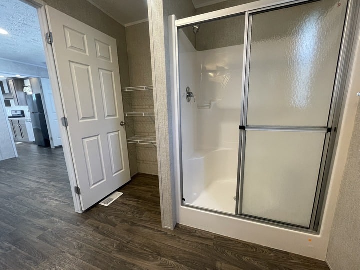 A bathroom with wood-look flooring, a glass shower enclosure, wire shelving, and an open door leading to a hallway and kitchen area.