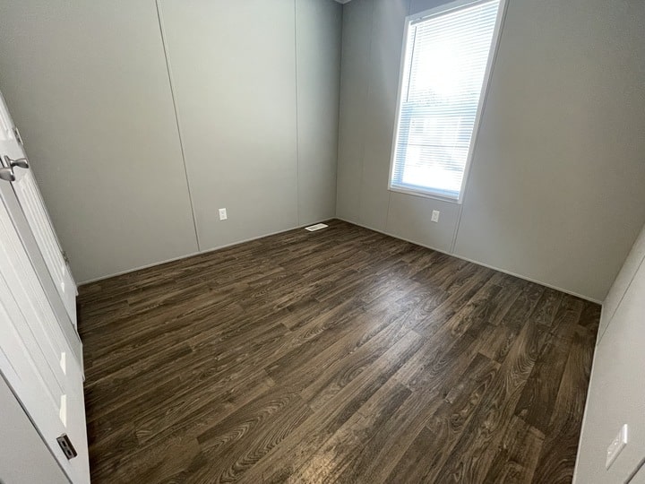 A small, empty room with light gray walls, dark wood flooring, a single window with blinds letting in daylight, and a closed white door to the left.