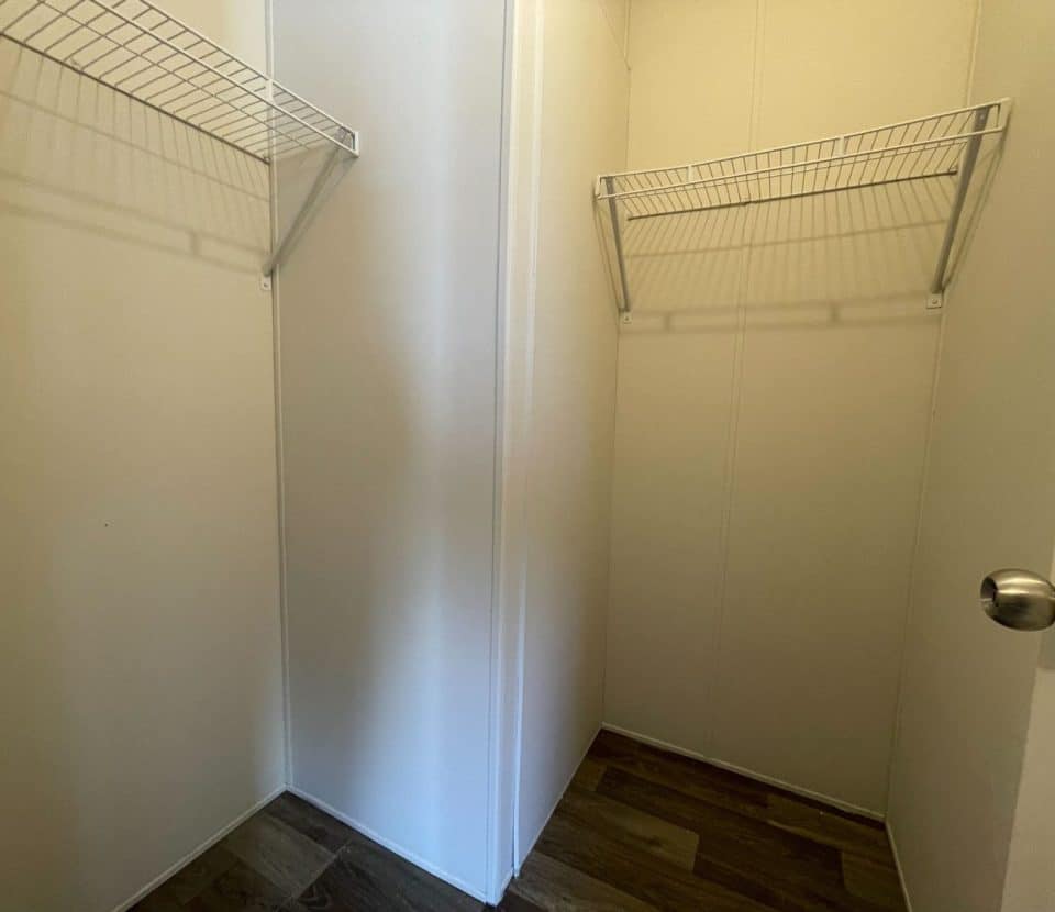 An empty walk-in closet with white walls, two wire shelves on opposite sides, a wooden floor, and a door with a round silver knob.