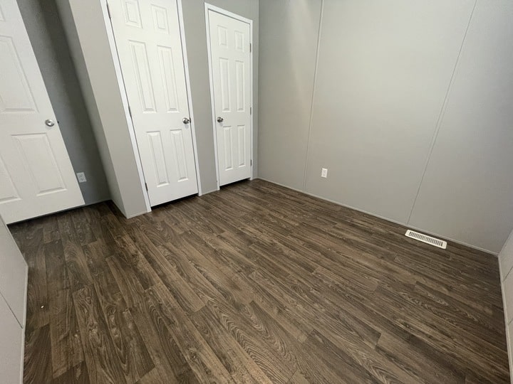 Empty room with light gray walls, dark wood flooring, and three white doors. There is a small white vent on the floor near the wall and a single electrical outlet visible.