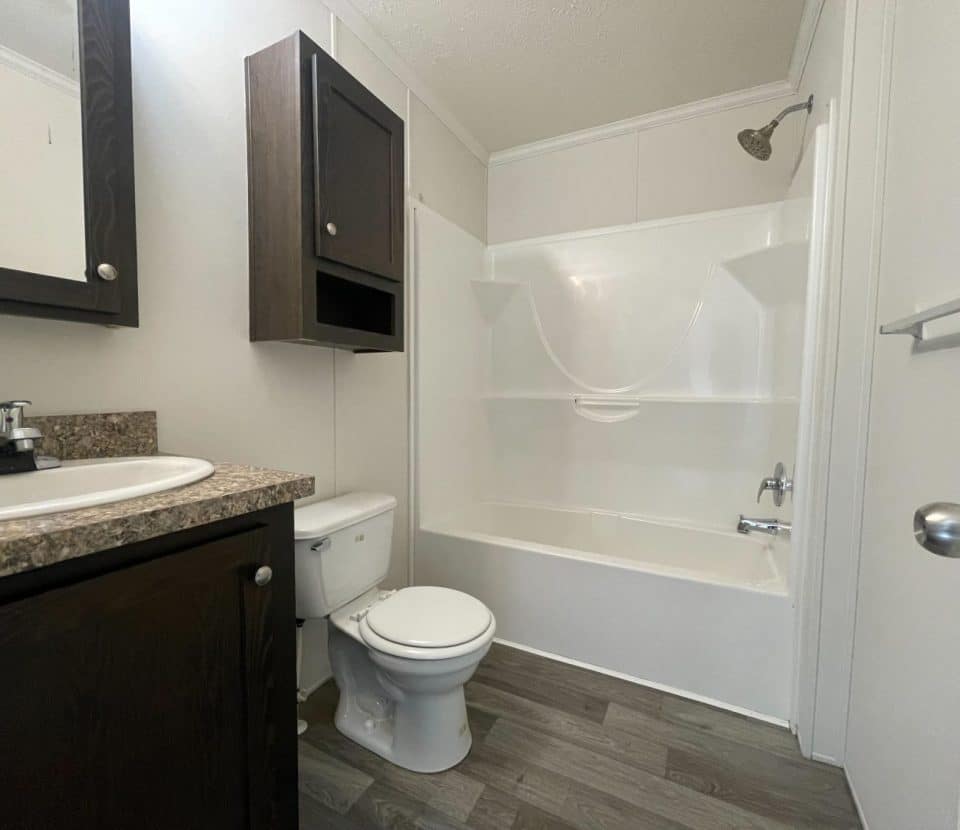 A small bathroom with a white bathtub and shower, a toilet, a sink with a granite countertop, dark wood cabinets, and gray wood-look flooring. The walls are light-colored, and the door is slightly open.