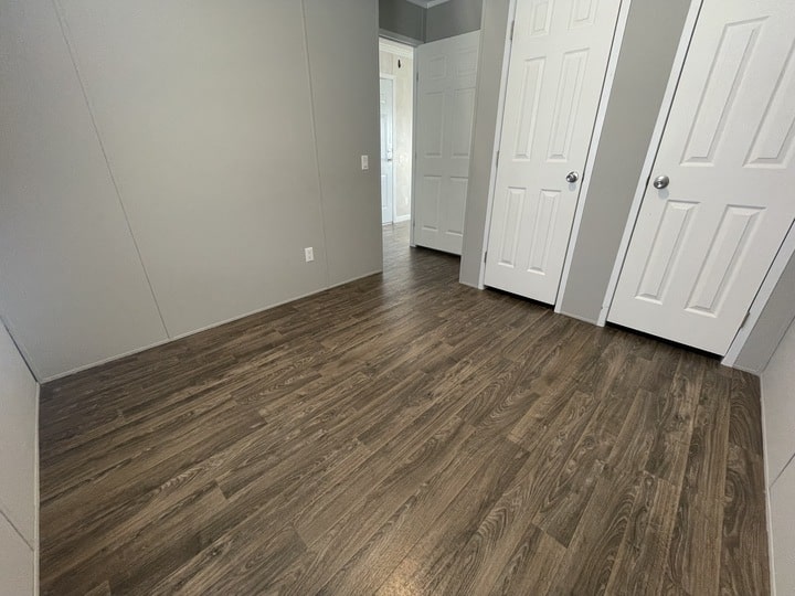 A small, empty room with gray walls, dark wood-look flooring, and three white doors—two on the right and one at the end of the room. The space is well-lit by natural light.