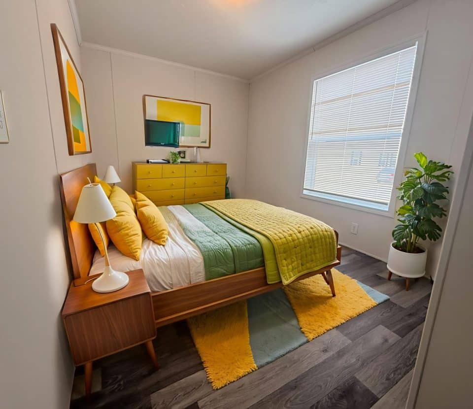 A modern bedroom with a wooden bed, yellow and green bedding, a matching dresser, a bedside lamp, wall art, a large window with blinds, a potted plant, and a yellow-green area rug on dark wood flooring.