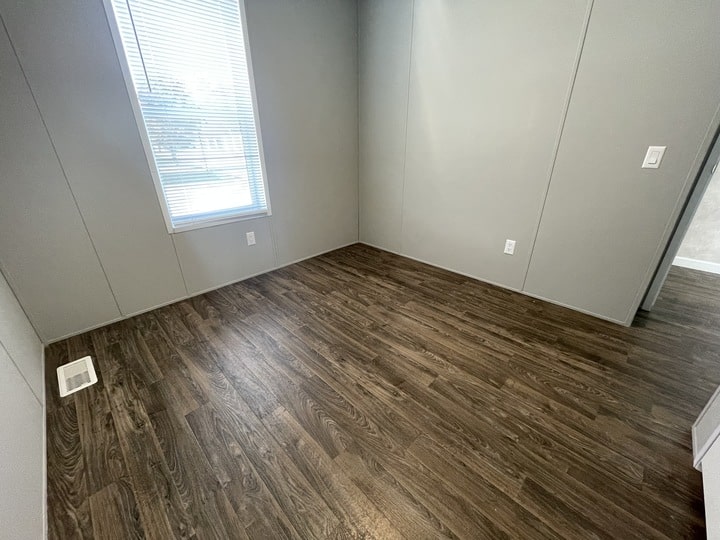 Small empty room with gray walls, a single large window letting in natural light, and dark wood-style flooring. An air vent is visible near the window, and there are electrical outlets on the walls.
