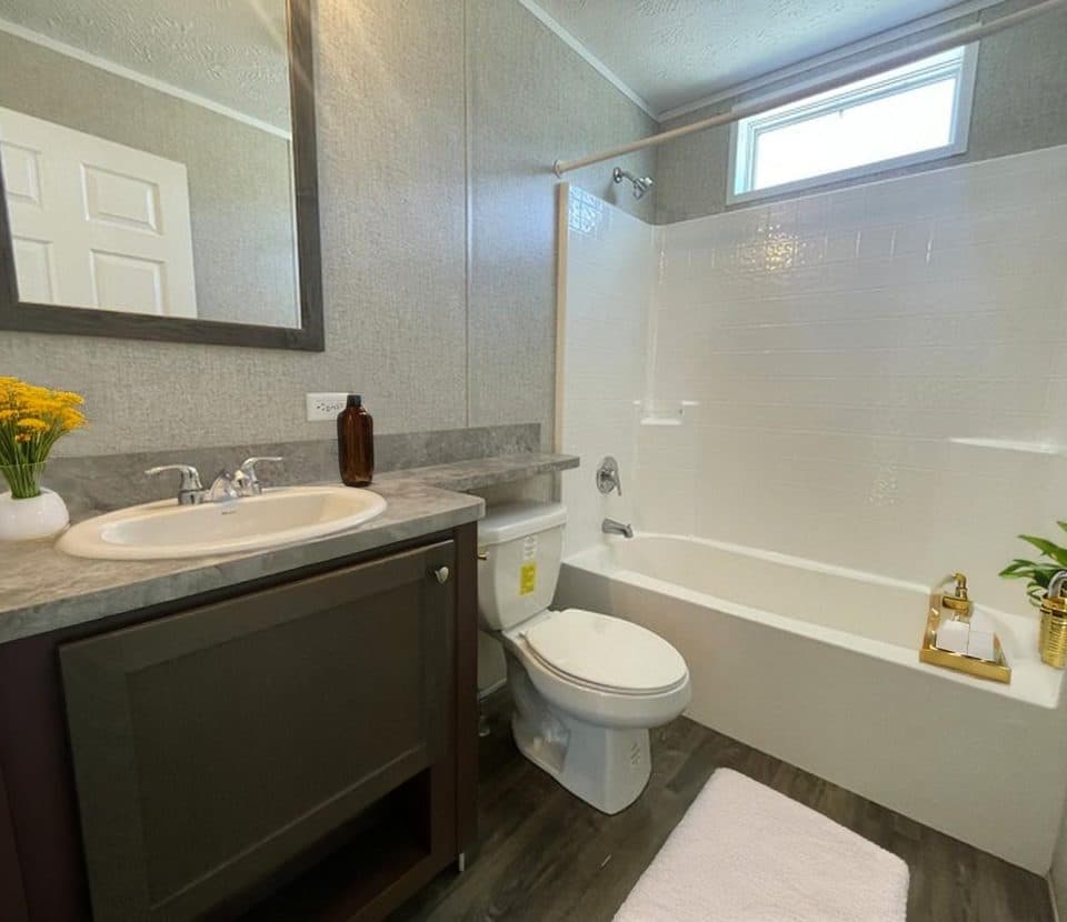 Modern bathroom with a sink and mirror on the left, toilet in the center, and a bathtub with a shower on the right. There are yellow flowers on the counter and a small plant on a tray in the tub.