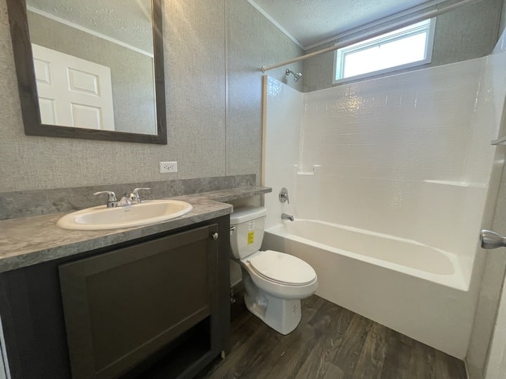 A small bathroom with a single sink vanity, a wall-mounted mirror, a toilet, and a white bathtub-shower combo under a high window. The floor is wood-patterned, and the countertop is gray.