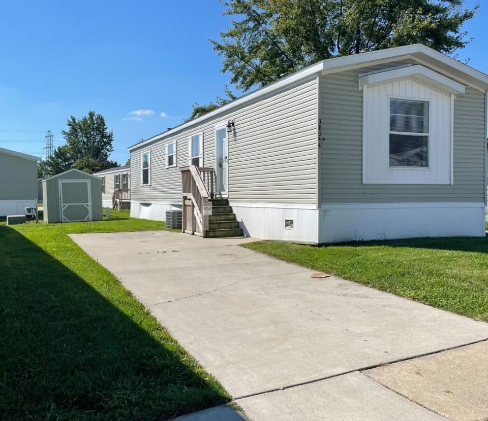 A light gray mobile home with white trim sits on a grassy lot, featuring a small porch, steps, a paved driveway, and a storage shed. The sky is clear and blue with trees in the background.