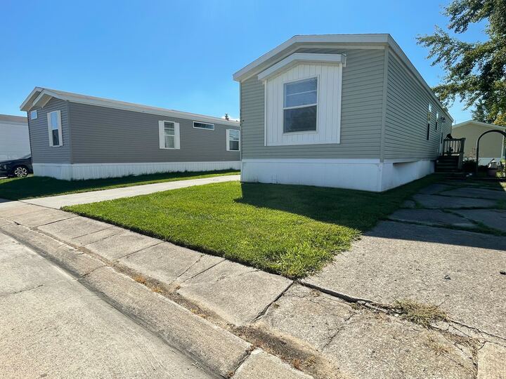 Two manufactured homes with gray siding and white trim sit side by side. One has a small grass lawn and driveway, while the other sits behind it. The sky is clear and sunny.