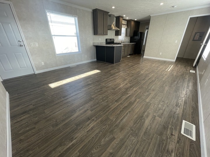 A spacious, empty room with dark wood flooring, light-colored walls, and a kitchen area featuring dark cabinets and stainless steel appliances in the background. Sunlight streams in from a window on the left.