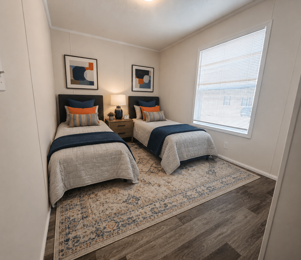 A bedroom with two twin beds, blue and white bedding, a nightstand with a lamp between them, framed abstract art above each bed, and a large window with blinds.