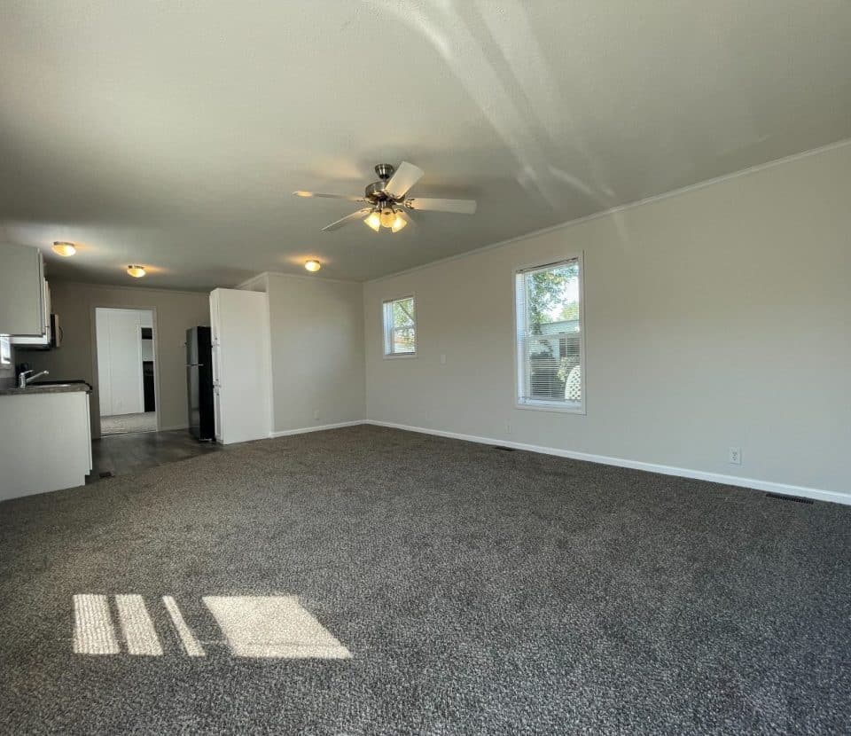 A spacious, unfurnished living room with gray carpet, light-colored walls, a ceiling fan with lights, and multiple windows letting in natural light. The kitchen area with appliances is visible in the background.