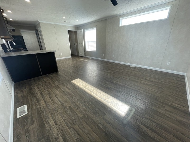 Spacious room with dark wood floors, beige walls, large window, and a small kitchen area with black cabinets on the left. Ceiling lights and natural light brighten the open space.