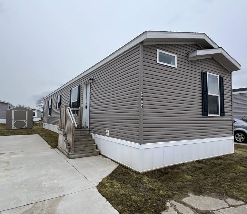 A gray manufactured home with white trim and a small wooden porch sits on a concrete driveway, with a car and another shed visible nearby under a cloudy sky.