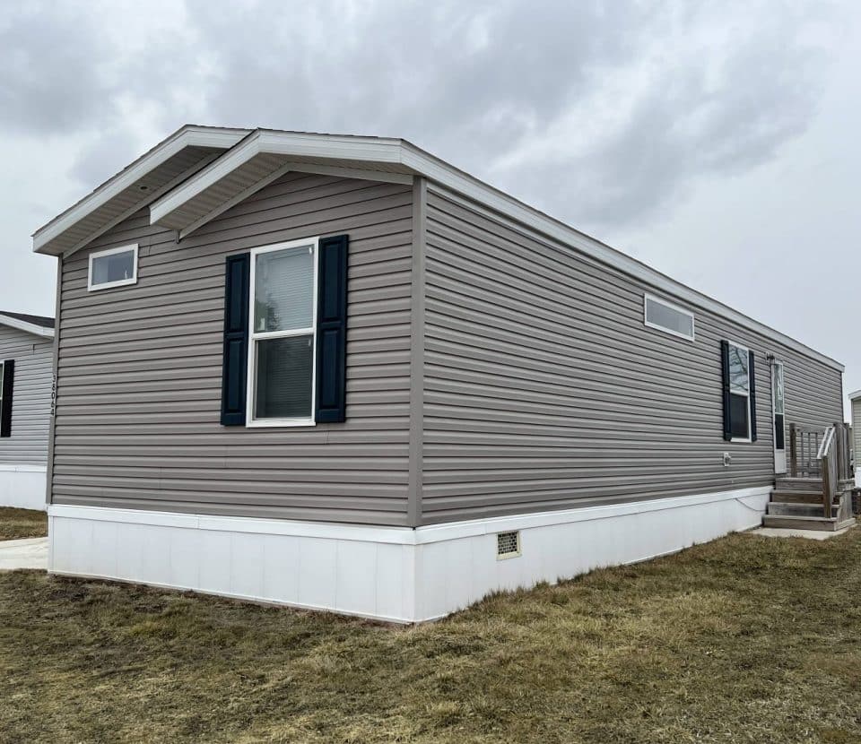 A beige manufactured home with white trim and dark shutters, set on a grassy lot with a small porch and steps leading to the entrance; overcast sky above.