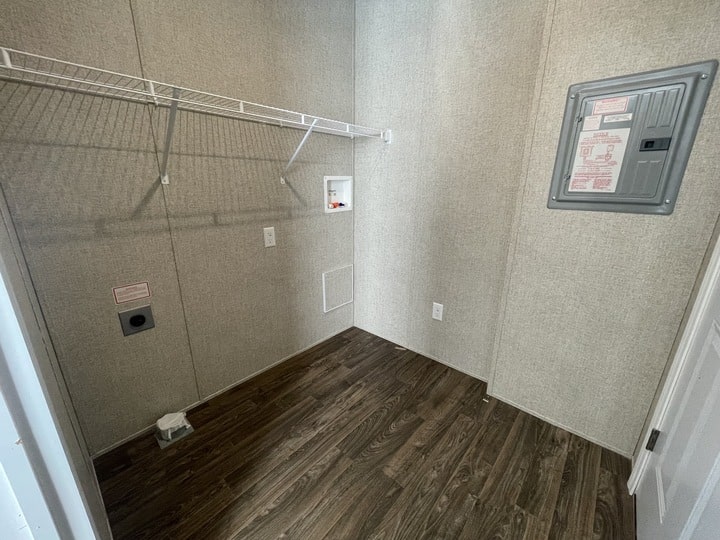 Empty laundry room with light-colored textured walls, a dark wood floor, a wire shelf, washer and dryer hookups, electrical outlets, and a gray electrical panel on the right wall.