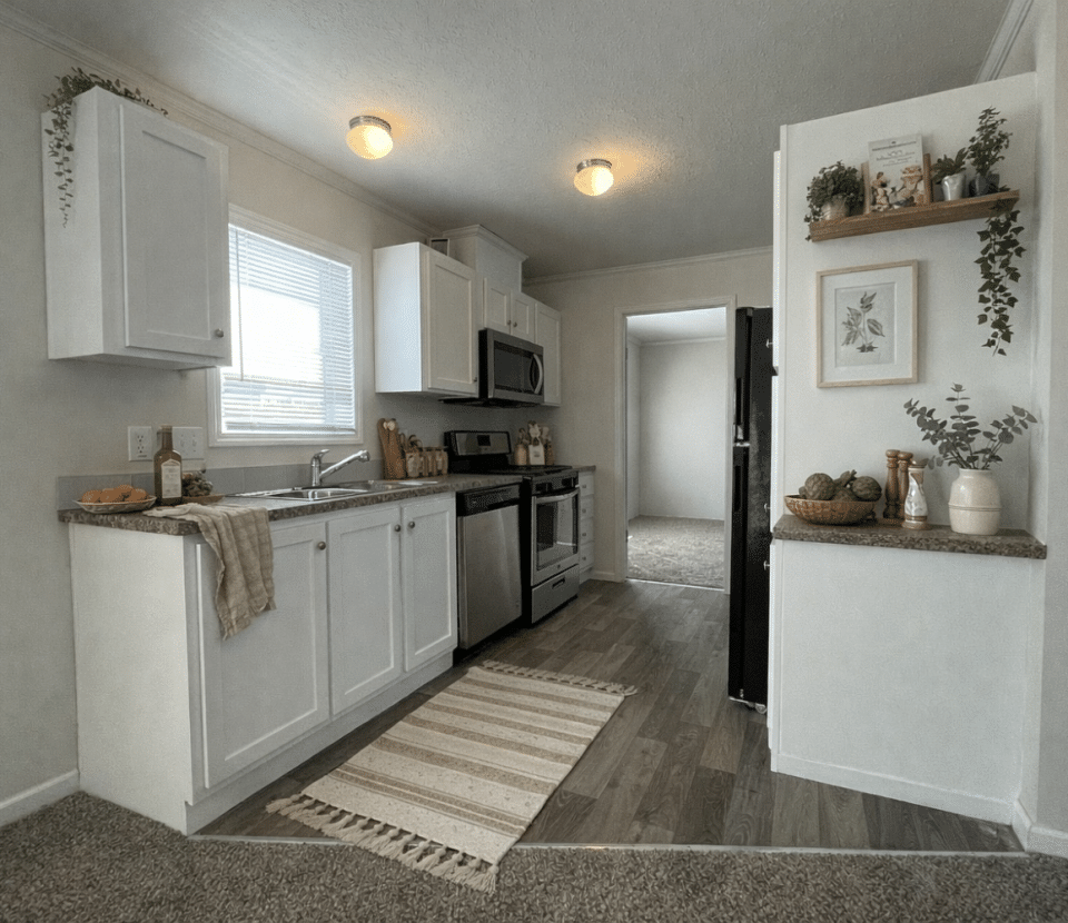 A modern kitchen with white cabinets, stainless steel appliances, a window above the sink, light wood flooring, and decorative plants on the countertops and shelves. A beige rug lies on the floor in front of the sink.