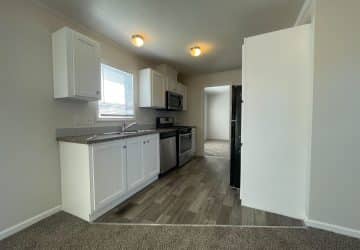 A small kitchen with white cabinets, stainless steel appliances, gray countertops, and laminate wood flooring, adjacent to a carpeted area and lit by two ceiling lights. A doorway leads to another room.