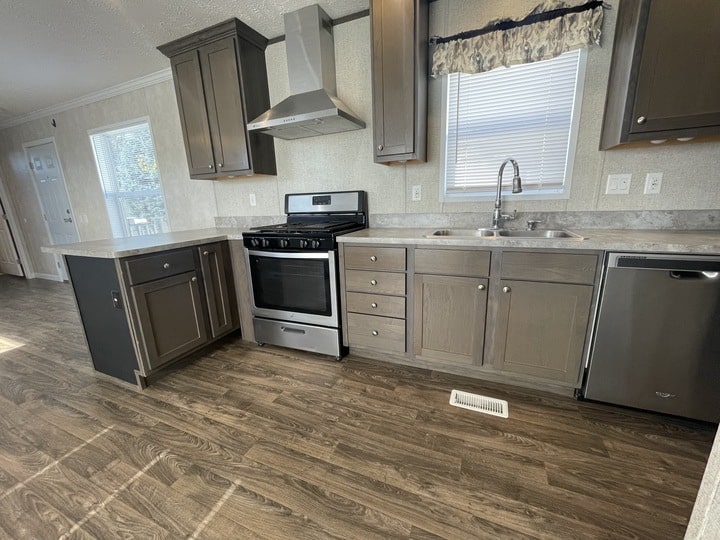 Modern kitchen with dark wood cabinets, stainless steel stove, oven hood, dishwasher, double sink, window with a valance, and light wood flooring. Countertops are a light gray color.