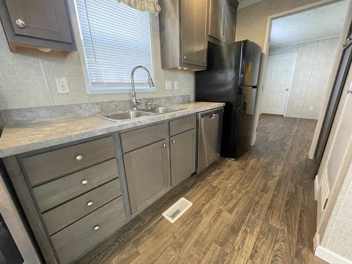 A modern kitchen with gray cabinets, stainless steel sink, dishwasher, black refrigerator, wood-style flooring, and a window above the sink letting in natural light.